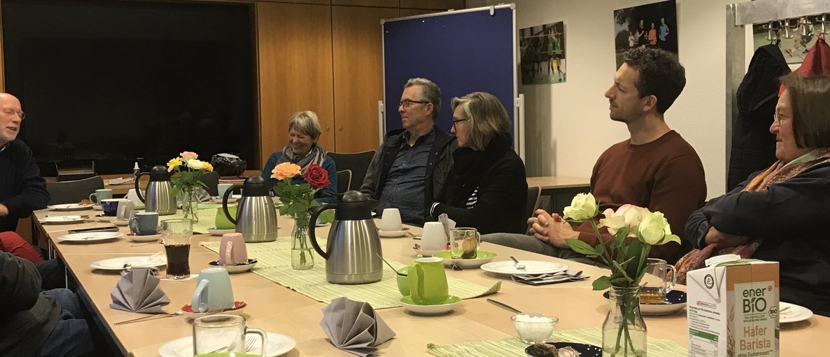 Mehrere Menschen sitzen an einem Kaffeetisch und hören einem Mann am Kopf des Tisches zu.